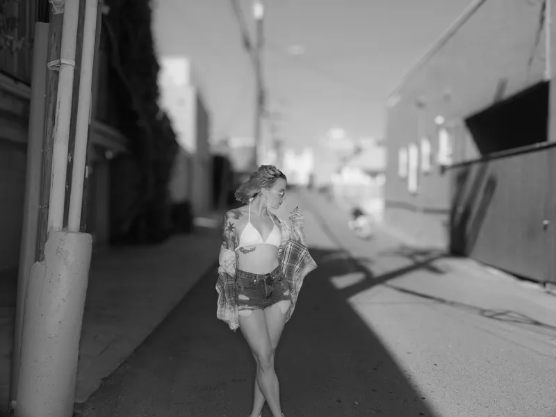 Tattooed woman in sunglasses holding a cigarette in a sunlit alley, wearing a bikini top, denim cutoffs and an open plaid shirt