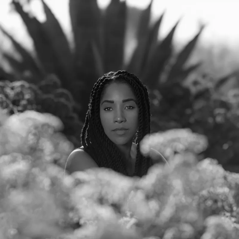 Ashley Blaine Featherson-Jenkins with long braids, framed by flowering shrubs and an agave plant