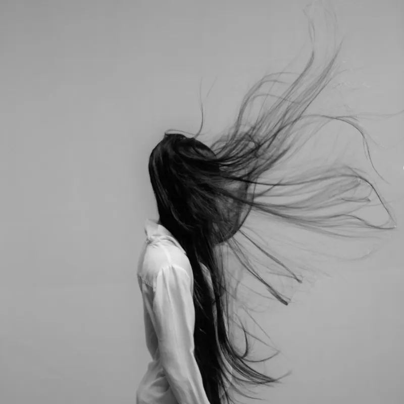 Woman in a white shirt with long dark hair flying across her face against a pale wall