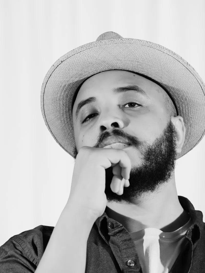 Justin Simien in a straw hat, chin resting on his hand, looking down at the camera