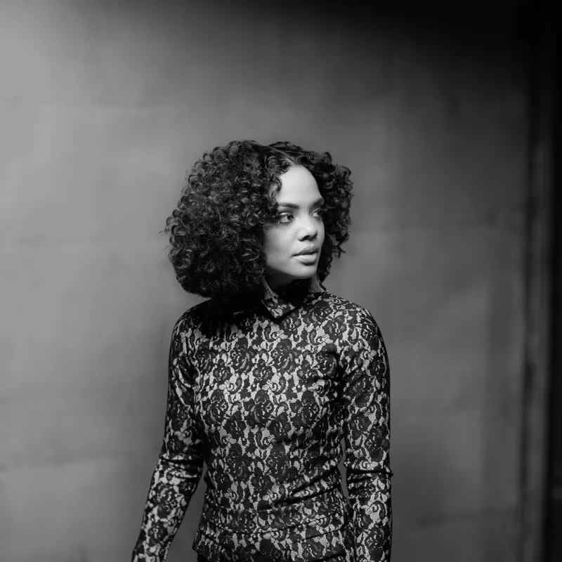 Woman with voluminous curls in a high-necked lace top, looking off to the side against a concrete wall