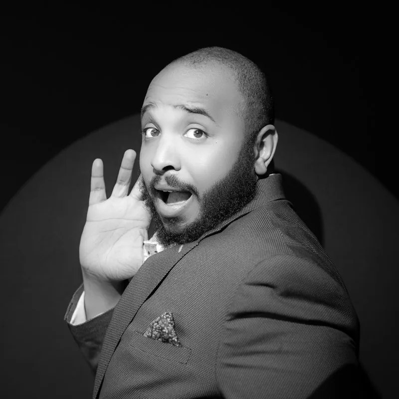 Bearded man in a suit and pocket square under a spotlight, mouth open in mock surprise, one hand raised