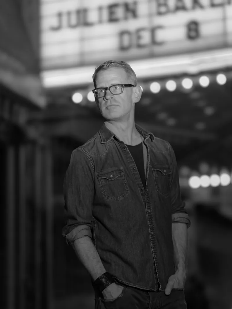 Man in glasses and a denim shirt standing beneath a lit theater marquee