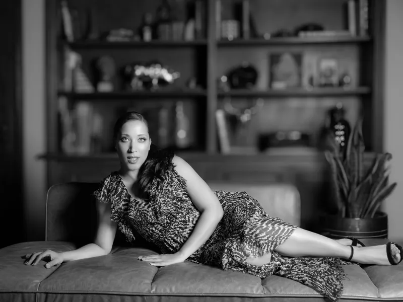 Robin Thede in a patterned dress, reclining on a leather daybed in front of bookshelves