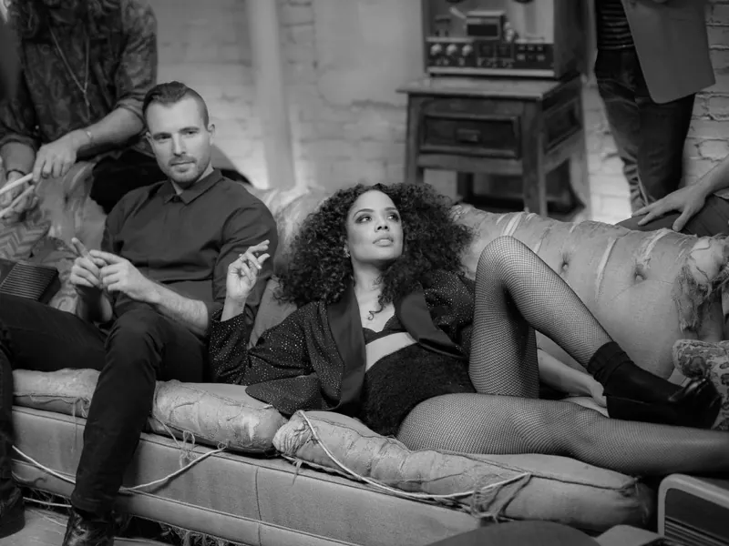 Tessa Thompson reclining on a worn sofa beside Jesse Nolan, with other people around them