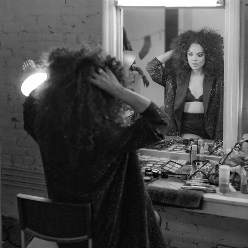 Tessa Thompson fixing her hair at a makeup mirror, her reflection facing the camera