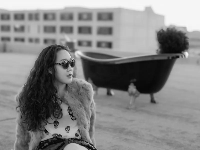 Woman with long curly hair and sunglasses in a fur coat on a rooftop, a clawfoot bathtub behind her