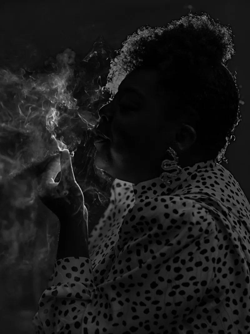 Woman in profile exhaling smoke in low light, wearing a spotted blouse and statement earrings