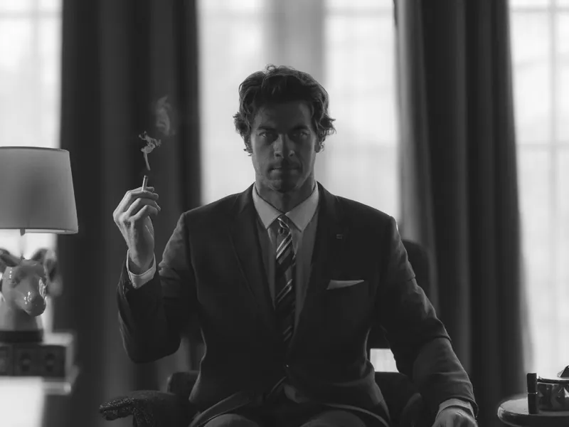 Man in a suit and tie seated in an armchair holding a cigarette, curtained windows behind him