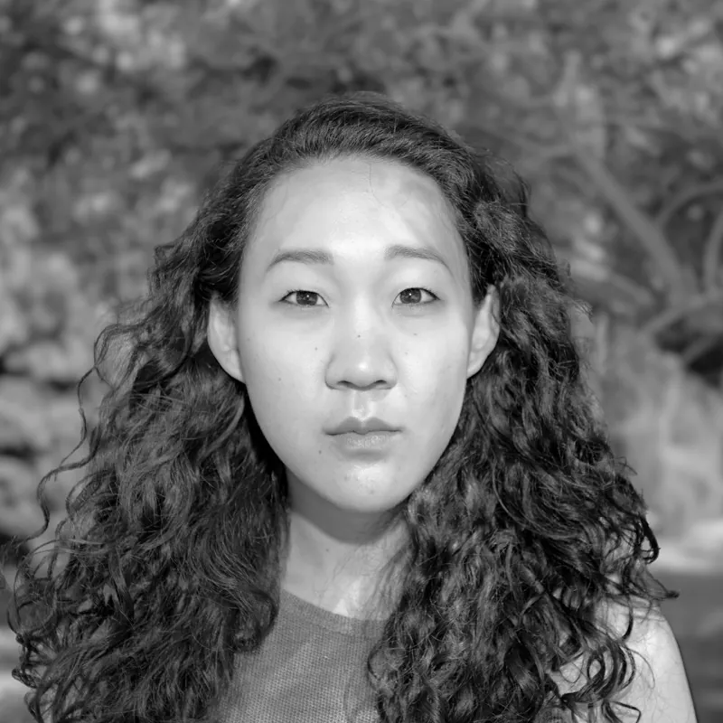 Woman with long curly hair looking straight at the camera, foliage behind her