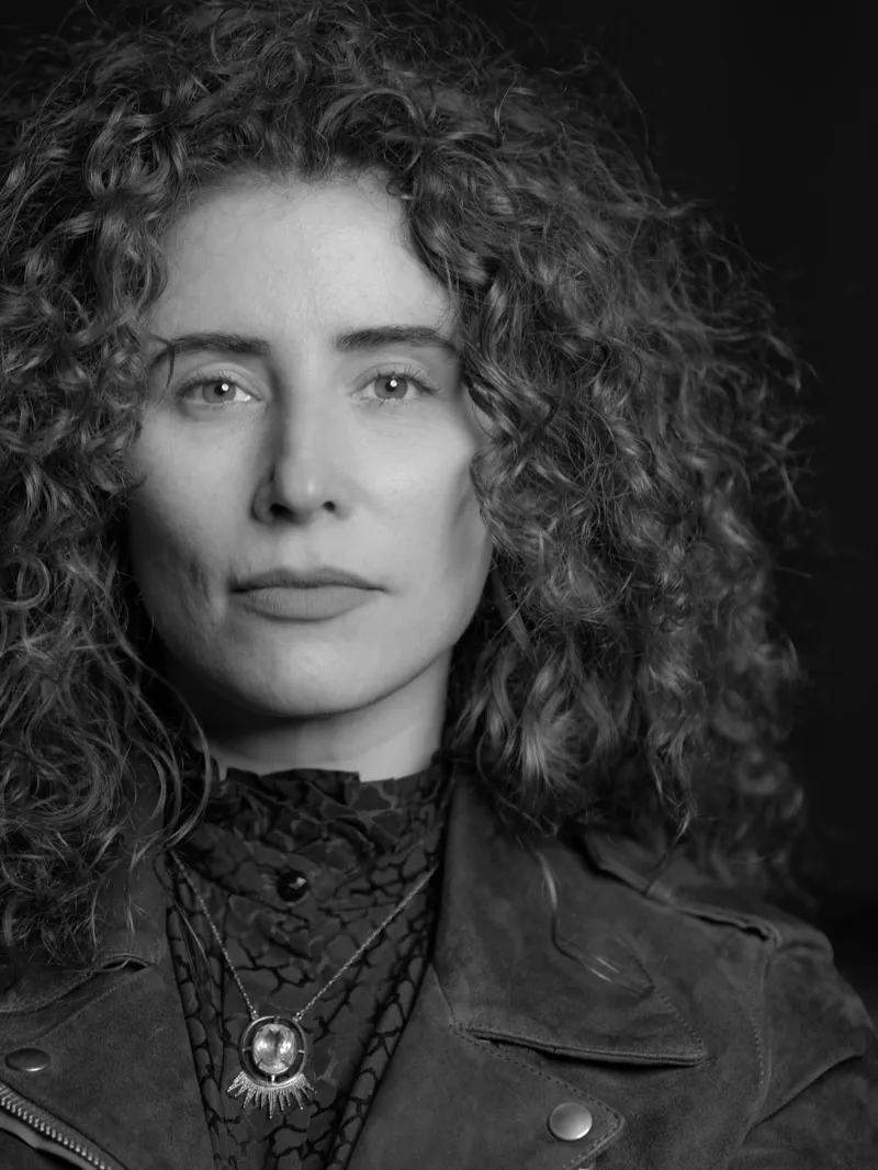 Woman with curly hair in a leather jacket and pendant necklace against a dark background