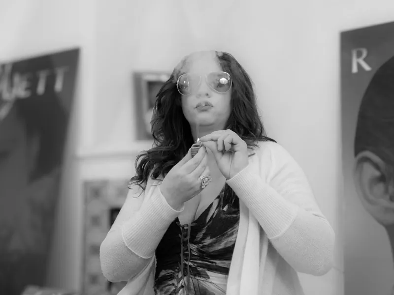 Woman in oversized round glasses lighting a cigarette in a cloud of smoke, paintings behind her