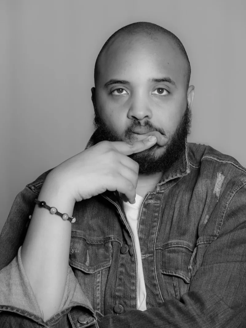 Bearded man in a denim jacket, finger resting on his lip, looking at the camera