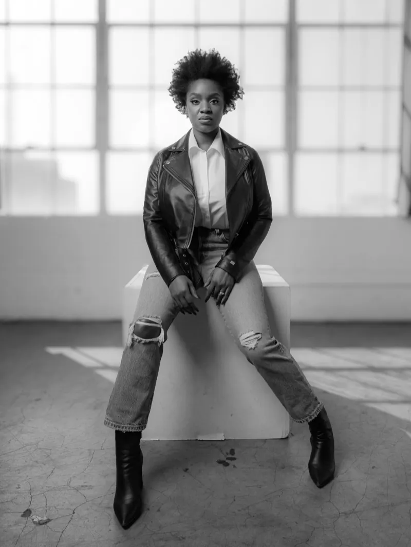 Woman in a leather jacket and ripped jeans seated on a white box in front of tall factory windows