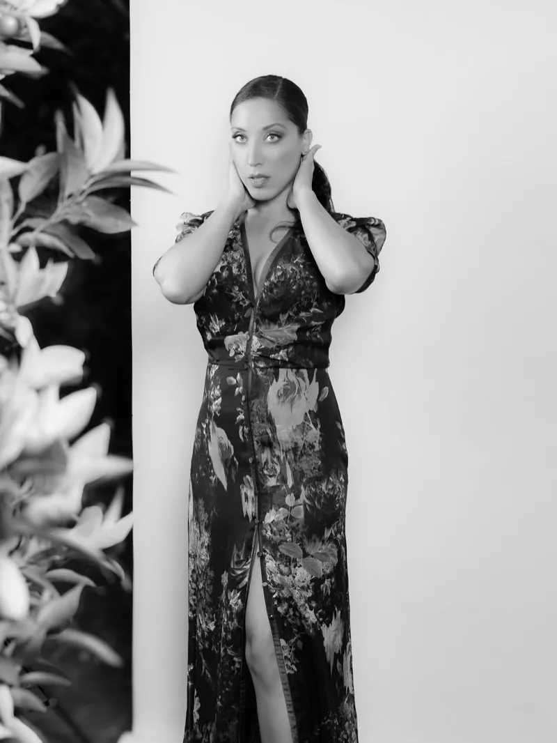 Woman in a long floral dress with her hands at her neck, standing beside leafy plants against a white wall