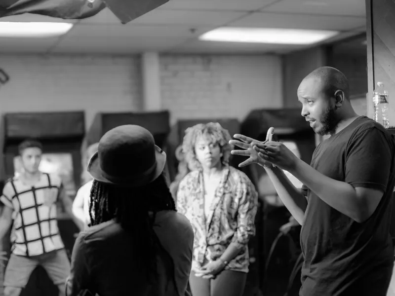 Man gesturing as he talks with a group of performers in a room lined with arcade cabinets