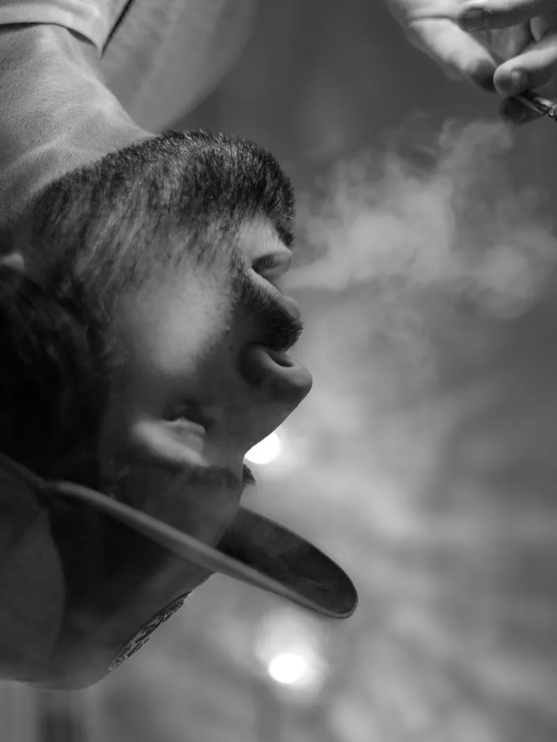 Upside-down close-up of a man in a baseball cap exhaling smoke, a hand holding a cigarette above him