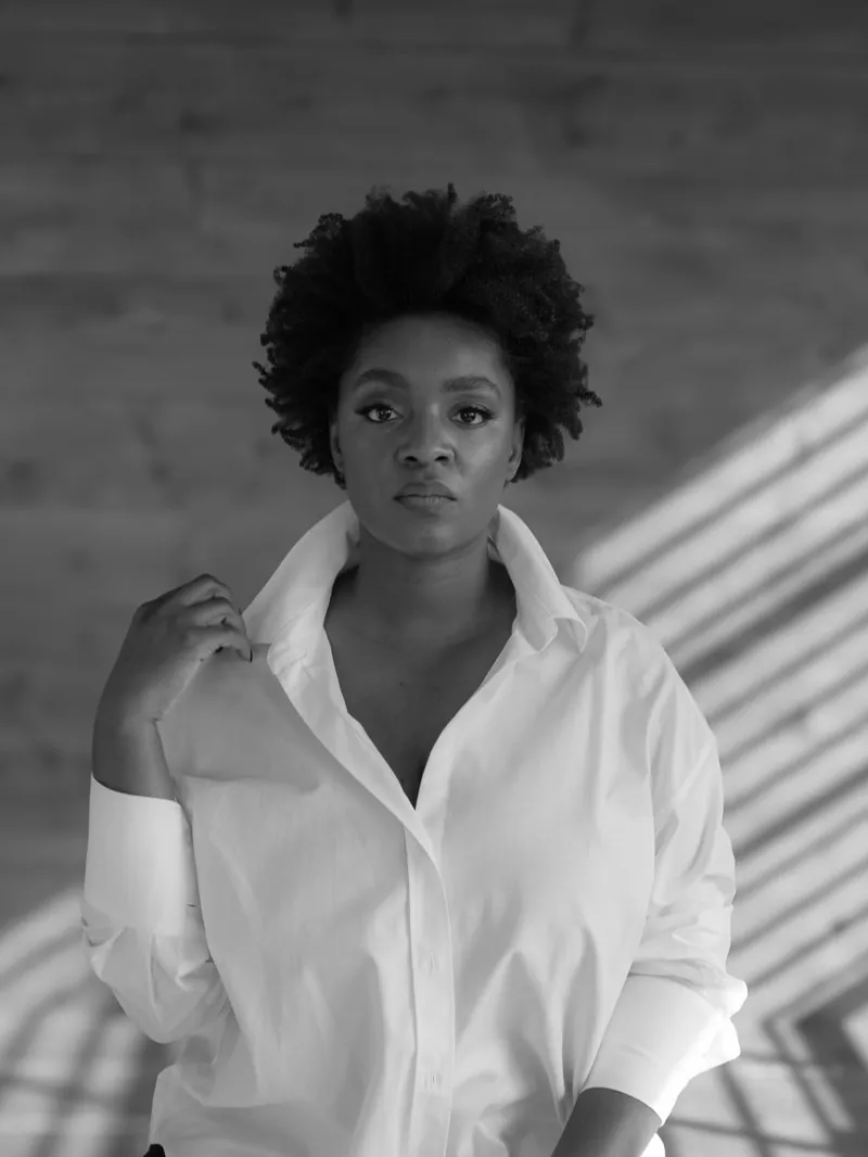Woman with natural hair in a white button-down shirt, window shadows on the wall behind her
